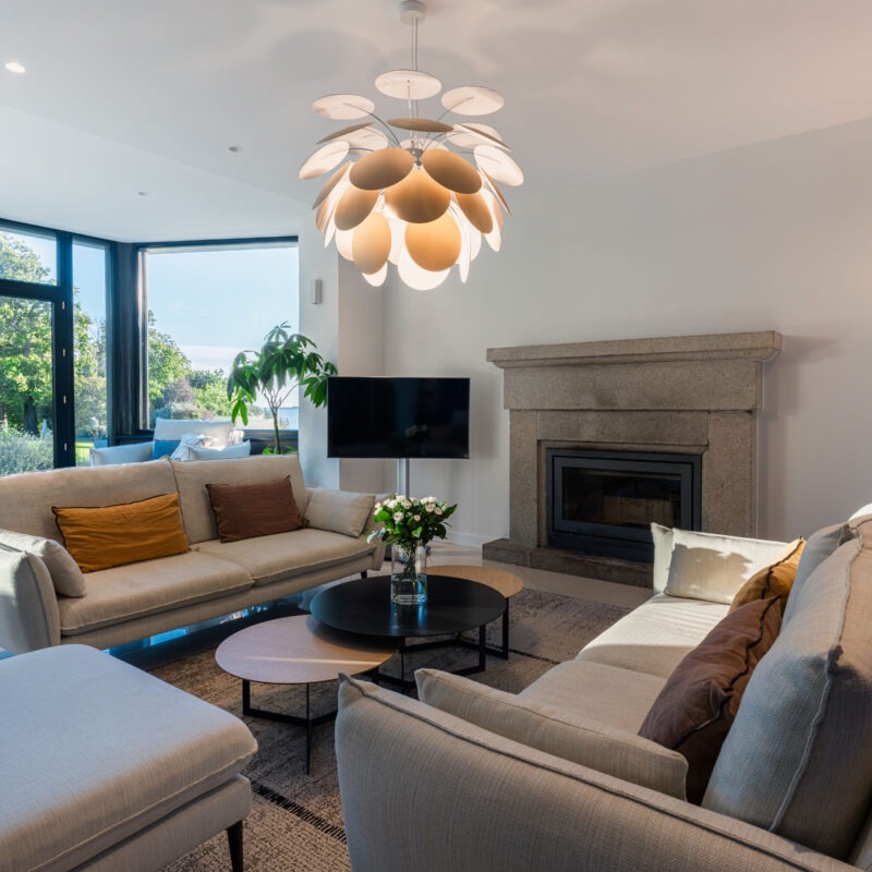 Salon moderne et lumineux avec grandes baies vitrées donnant sur un jardin arboré, cheminée en pierre et canapé beige