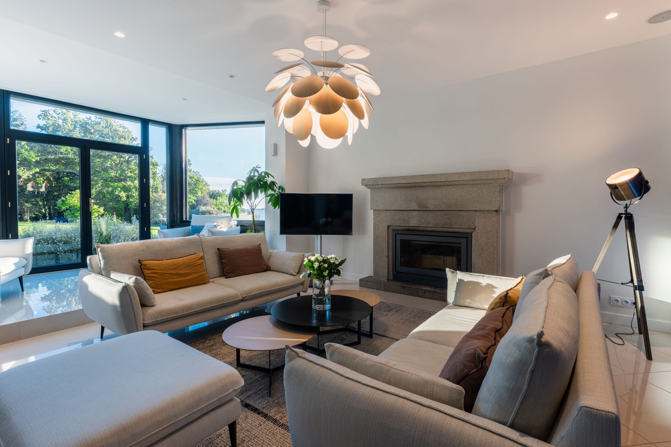 Salon moderne et lumineux avec grandes baies vitrées donnant sur un jardin arboré, cheminée en pierre et canapé beige