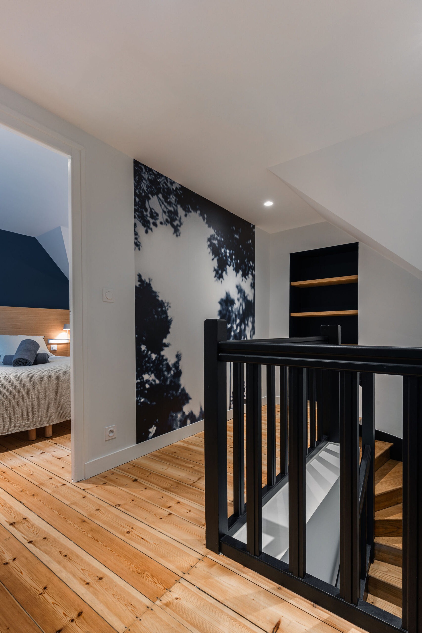 Palier avec escalier en bois et rampe noire, mur décoratif artistique et vue sur une chambre avec lit double