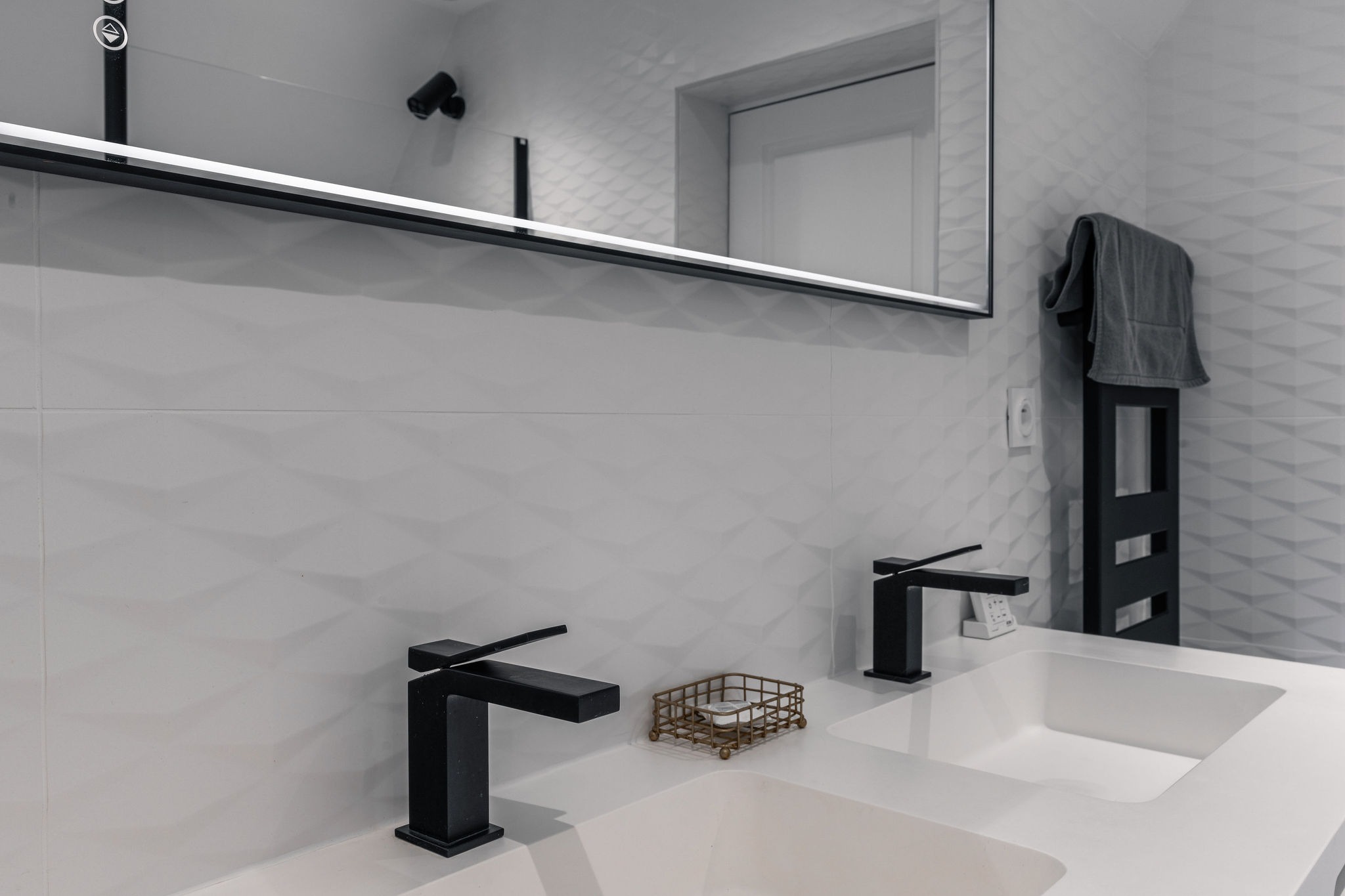 Double vasque blanche avec robinets noirs modernes, miroir rétroéclairé et mur en carrelage texturé – salle de bain du Domaine de Stang Bihan