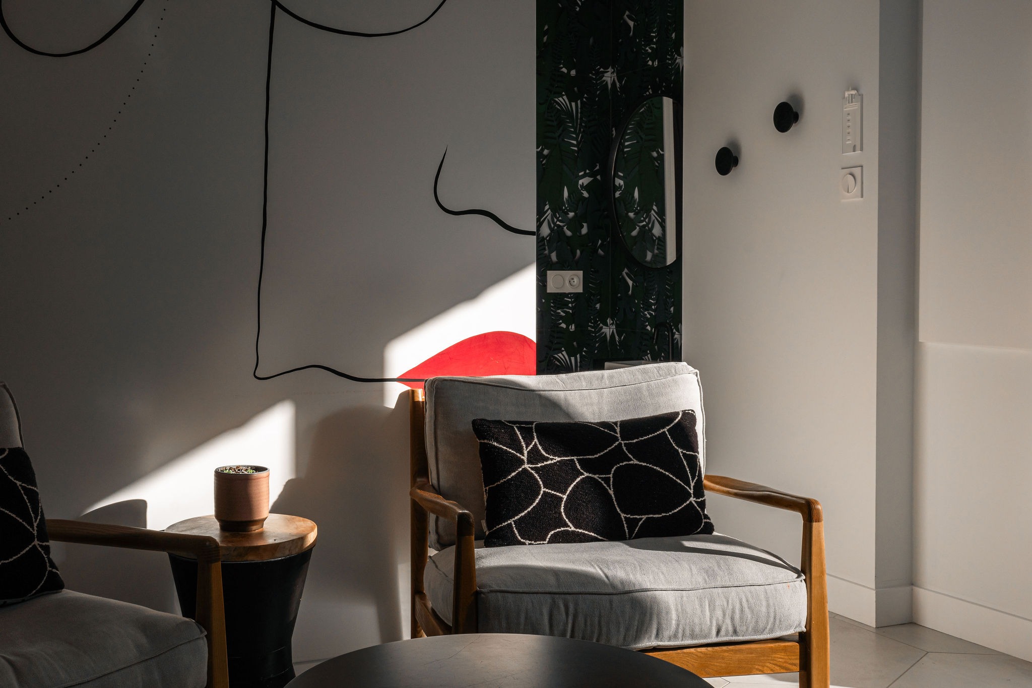 Détail d’un fauteuil en bois et tissu gris dans le salon, éclairé par le soleil du matin, avec coussin graphique et fresque murale artistique
