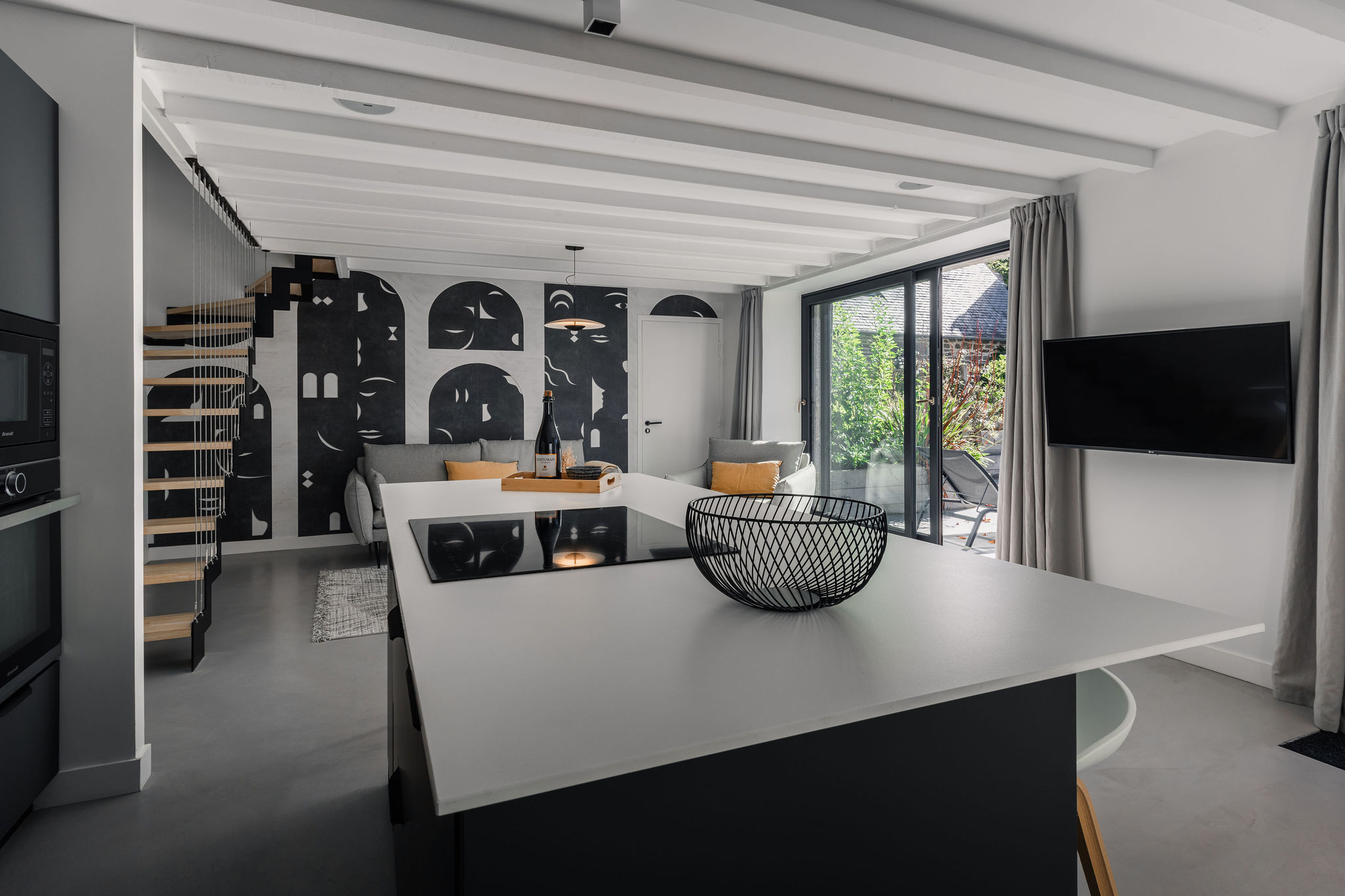 Vue de la cuisine ouverte et du salon moderne du logement Belem, avec escalier suspendu et grandes baies vitrées donnant sur la terrasse, dans une dépendance du Manoir de Stang Bihan.