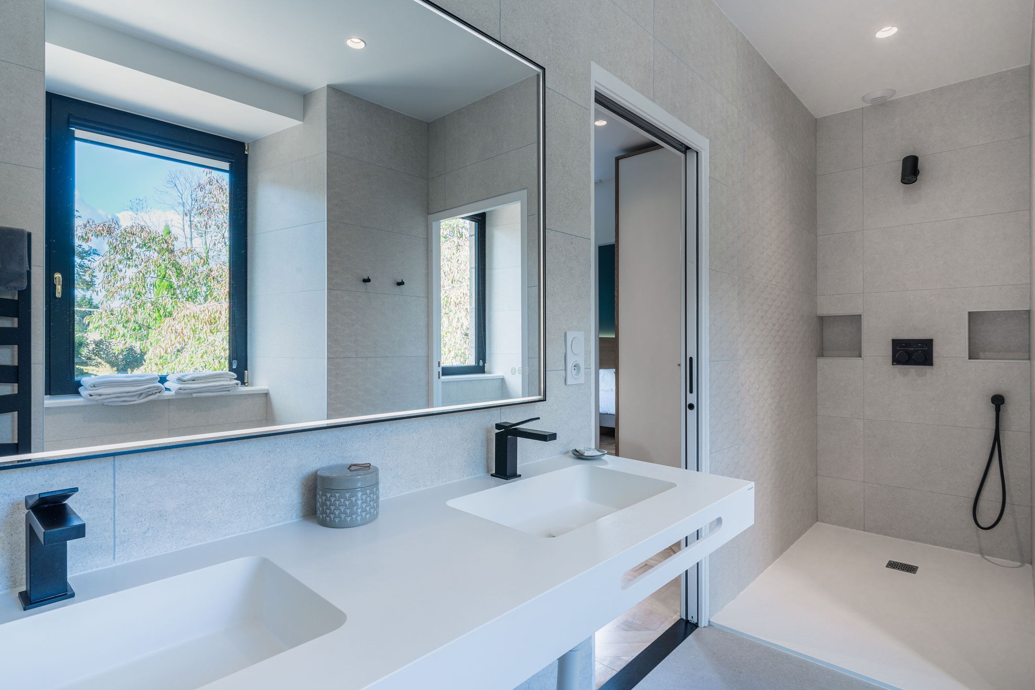 Salle de bain moderne de l’appartement La Fée de l’Aulne, avec double vasque, grande douche à l’italienne, robinetterie noire et vue sur la nature