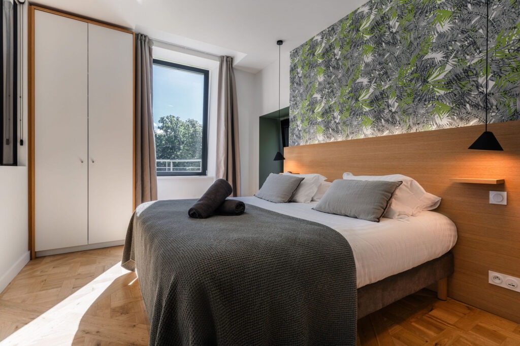 Chambre parentale avec grand lit double, lumière naturelle et décoration murale tropicale – Appartement Le Biche, Domaine de Stang Bihan à Concarneau